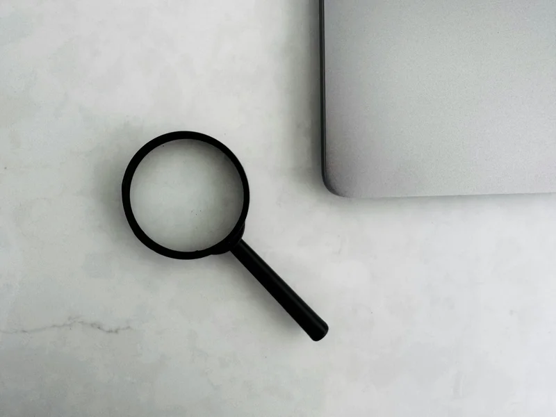A black magnifying glass is positioned next to a silver laptop on a light-colored surface. The image highlights the magnifying glass as a tool for search and examination, suggesting focus and inquiry.