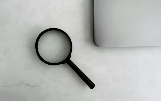 A black magnifying glass is positioned next to a silver laptop on a light-colored surface. The image highlights the magnifying glass as a tool for search and examination, suggesting focus and inquiry.