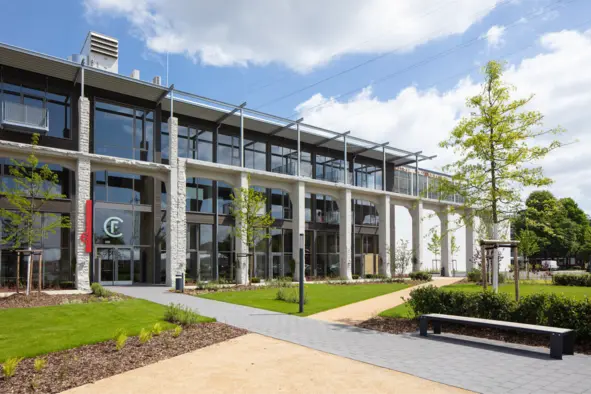 The arcade building on the Factory Campus in Düsseldorf features large windows and an entrance area. In front of the building stretches a well-kept green area with small trees and a path made of gray paving stones. A bench is also visible. The sky is partly cloudy.
