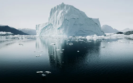 A towering, jagged iceberg rises from calm dark water, surrounded by scattered chunks of ice. The massive white structure is reflected in the rippling surface, with distant mountains visible in the background.