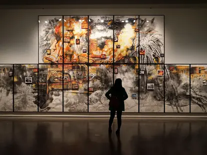 A person stands in front of a large, vibrant artwork consisting of multiple panels. The artwork features bold colors like orange and black, depicting abstract forms and smaller framed images scattered throughout. The scene captures the viewer's engagement with the artistic display.